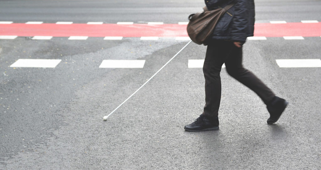 Dieses Bild zeigt den Unterkörper einer Person die eine Straße überquert und einen Blindenstock in der Hand hält